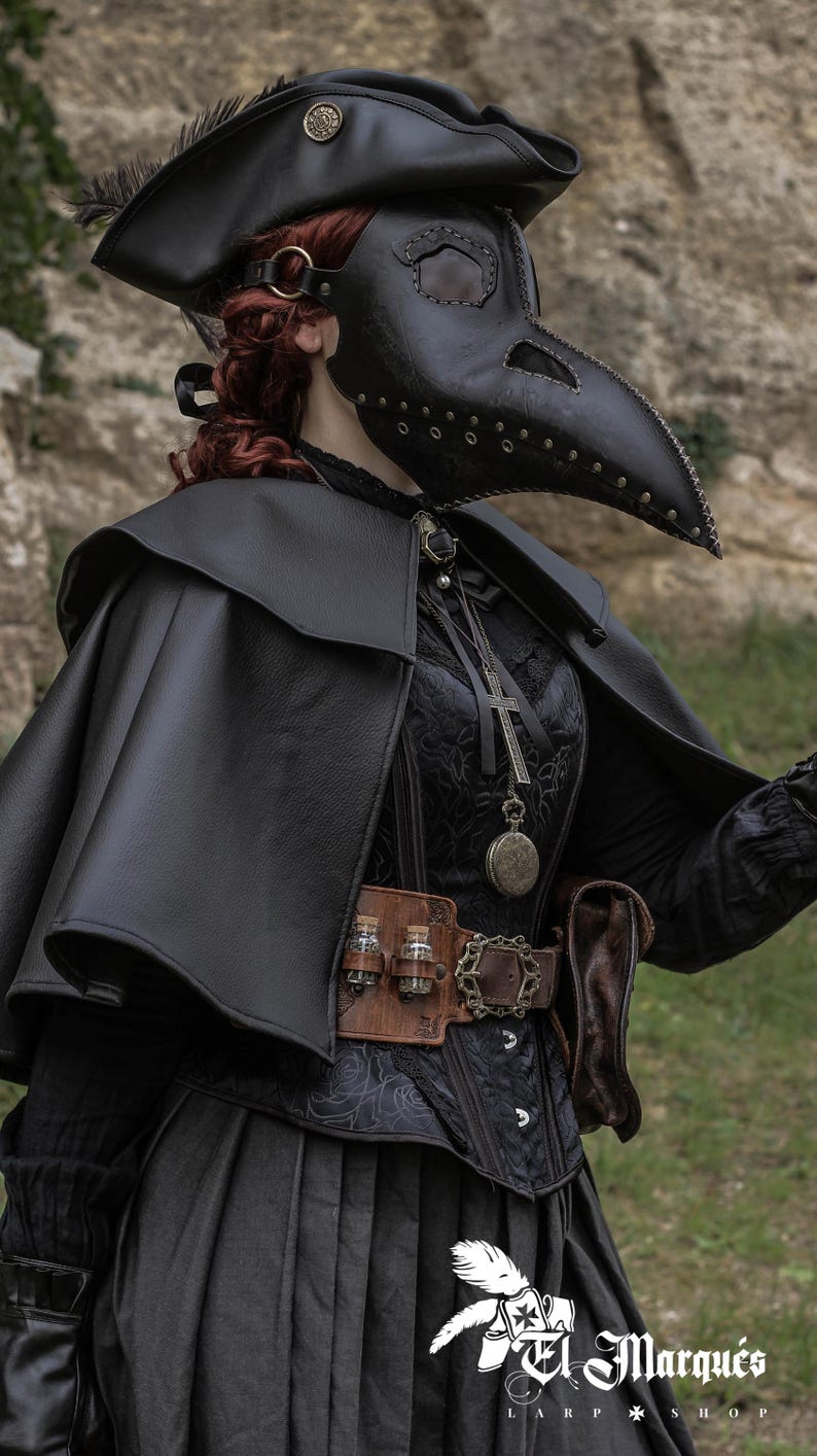 May include: A person wearing a black leather plague doctor mask with a long beak, a black leather hat, a black cape, and a black dress. The person is holding a gun in their right hand. The background is a rocky wall.