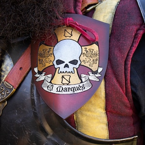 K&ouml;nnte beinhalten: Ein roter, goldener und wei&szlig;er Schild mit einem Totenkopf- und Knochenkreuz-Design. Der Schild tr&auml;gt den Text "El Marques".