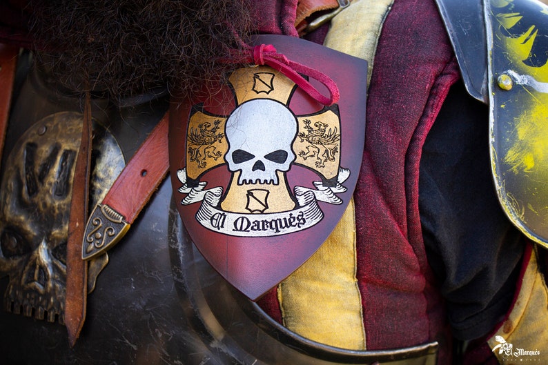 K&ouml;nnte beinhalten: Ein roter, goldener und wei&szlig;er Schild mit einem Totenkopf- und Knochenkreuz-Design. Der Schild tr&auml;gt den Text "El Marques".