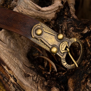 May include: A brown leather belt with a brass buckle featuring a Celtic knot design. The buckle has a decorative, ornate design with three round accents.