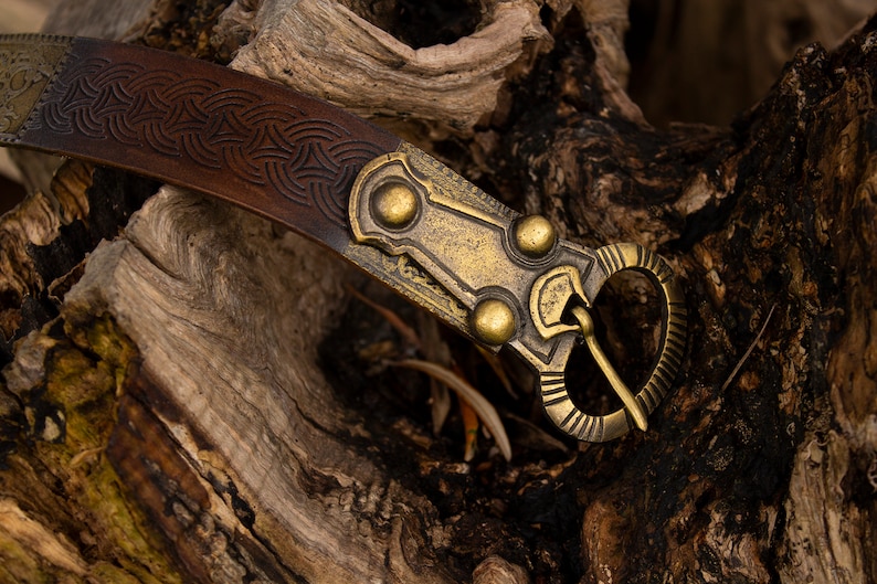 May include: A brown leather belt with a brass buckle featuring a Celtic knot design. The buckle has a decorative, ornate design with three round accents.