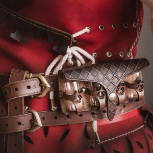 Peut inclure: Gros plan sur un v&ecirc;tement rouge avec une ceinture en cuir marron et une petite pochette. La ceinture a une boucle en laiton et la pochette a un motif en losanges. Le v&ecirc;tement a des &oelig;illets et des lacets blancs.