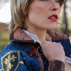 K&ouml;nnte beinhalten: Eine Frau in einem mittelalterlichen Kost&uuml;m in Blau und Braun mit einem goldenen Fleur-de-lis-Emblem am Arm. Sie h&auml;lt ein Schwert in ihrer rechten Hand und tr&auml;gt ein Lederarmband mit einem Fleur-de-lis-Design am linken Handgelenk.
