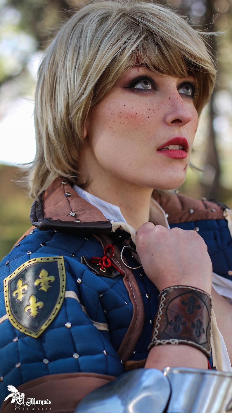 K&ouml;nnte beinhalten: Eine Frau in einem mittelalterlichen Kost&uuml;m in Blau und Braun mit einem goldenen Fleur-de-lis-Emblem am Arm. Sie h&auml;lt ein Schwert in ihrer rechten Hand und tr&auml;gt ein Lederarmband mit einem Fleur-de-lis-Design am linken Handgelenk.