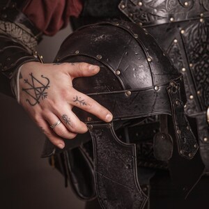 K&ouml;nnte beinhalten: Ein schwarzer, verwitterter Metallhelm mit Nietendetails, vor dunkler Kleidung und R&uuml;stung. Der Helm hat ein Visier und ist Teil eines mittelalterlichen oder Fantasy-Ensembles. Das Bild suggeriert ein historisches oder fiktives Kampfthema.