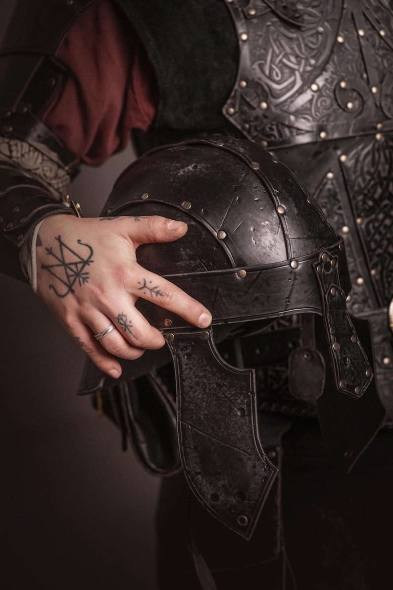 K&ouml;nnte beinhalten: Ein schwarzer, verwitterter Metallhelm mit Nietendetails, vor dunkler Kleidung und R&uuml;stung. Der Helm hat ein Visier und ist Teil eines mittelalterlichen oder Fantasy-Ensembles. Das Bild suggeriert ein historisches oder fiktives Kampfthema.