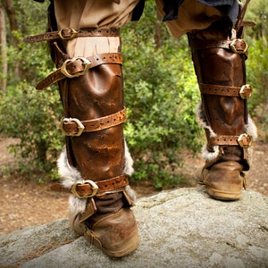 May include: Brown leather leg armor with gold buckles and fur trim. The armor is worn over brown boots.