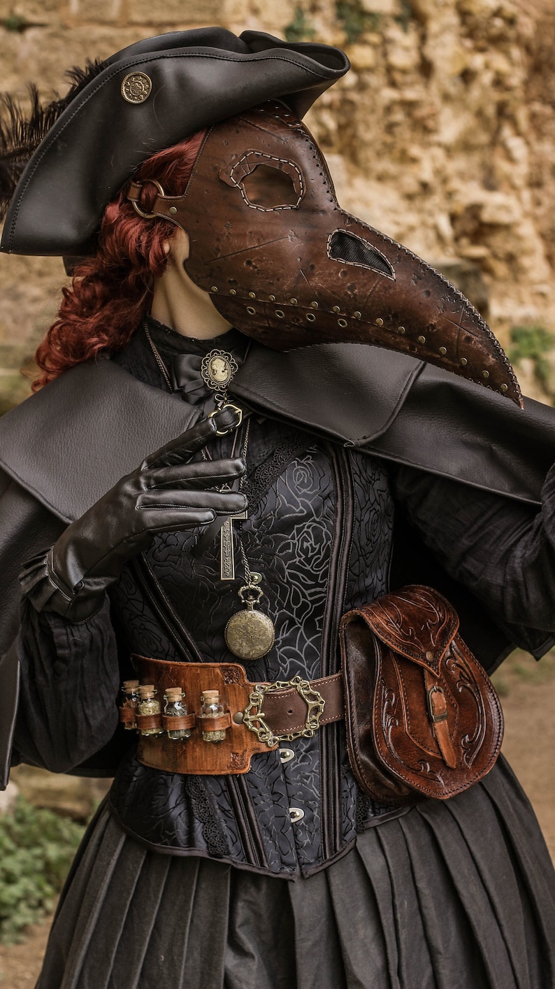 May include: A woman wearing a black and brown plague doctor costume with a long, pointed beak mask. She is holding a cross pendant and has a leather belt with vials and a pouch.