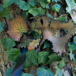 May include: A leather masquerade mask with a leafy design in shades of brown, orange, and green. The mask is attached to a black ribbon and is resting against a tree trunk covered in ivy.