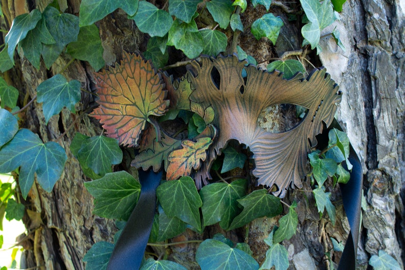 May include: A leather masquerade mask with a leafy design in shades of brown, orange, and green. The mask is attached to a black ribbon and is resting against a tree trunk covered in ivy.