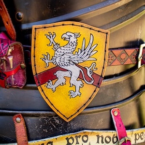 K&ouml;nnte beinhalten: Ein gelber Lederschild mit einem wei&szlig;en Greifen-Design und einem roten Streifen in der Mitte. Der Schild ist an einem braunen Lederg&uuml;rtel mit Totenkopf- und Knochenkreuz-Design befestigt.