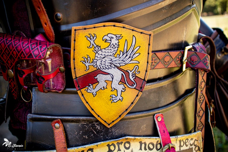 K&ouml;nnte beinhalten: Ein gelber Lederschild mit einem wei&szlig;en Greifen-Design und einem roten Streifen in der Mitte. Der Schild ist an einem braunen Lederg&uuml;rtel mit Totenkopf- und Knochenkreuz-Design befestigt.