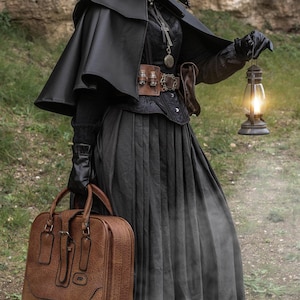 May include: A woman wearing a black plague doctor mask and a black cloak holds a lantern and a brown leather suitcase. She is standing in front of a rocky wall.