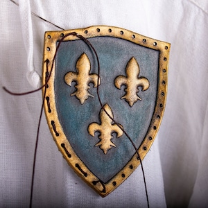 K&ouml;nnte beinhalten: Nahaufnahme eines Lederschildes mit drei goldenen Lilien auf blauem Hintergrund. Der Schild wird mit braunem Faden an einen wei&szlig;en Stoff gen&auml;ht.