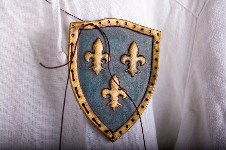 K&ouml;nnte beinhalten: Nahaufnahme eines Lederschildes mit drei goldenen Lilien auf blauem Hintergrund. Der Schild wird mit braunem Faden an einen wei&szlig;en Stoff gen&auml;ht.