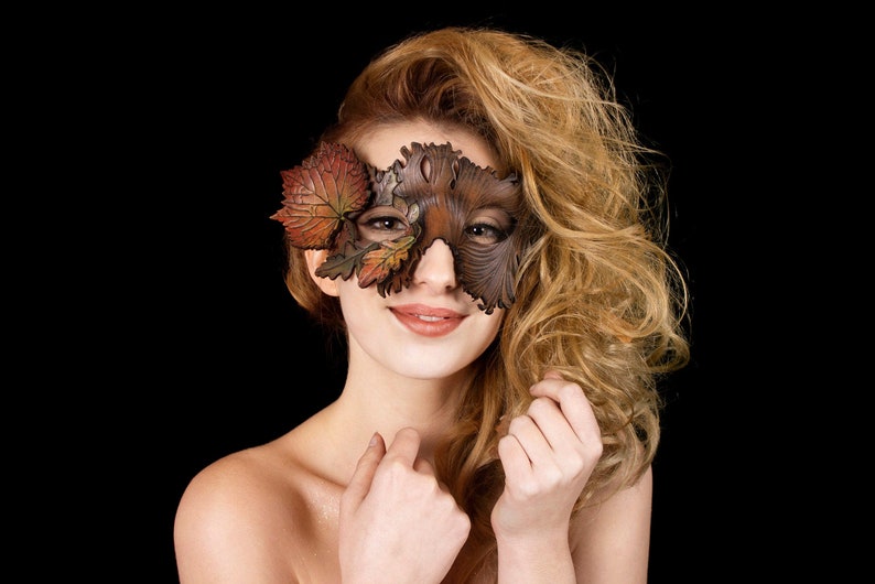 May include: A woman with long blonde hair wears a brown leather mask with a leaf design. The mask covers her eyes and part of her forehead. She is smiling and looking at the camera.