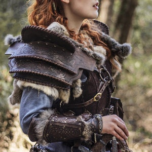 May include: A woman wearing a brown leather and fur costume with intricate designs. She is holding a sword in a leather scabbard. The costume includes a leather breastplate, shoulder pads, and arm guards. The woman has red hair and blue face paint.