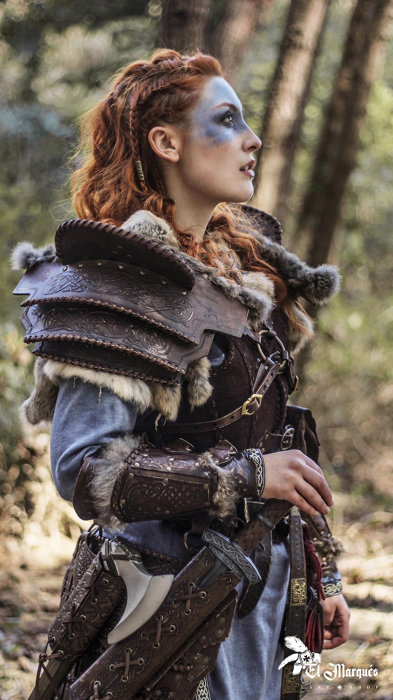 May include: A woman wearing a brown leather and fur costume with intricate designs. She is holding a sword in a leather scabbard. The costume includes a leather breastplate, shoulder pads, and arm guards. The woman has red hair and blue face paint.