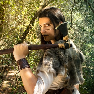 May include: A woman with dark hair and dark makeup is holding a small axe over her shoulder. She is wearing a fur-trimmed garment and a leather wristband. The background is a forest.