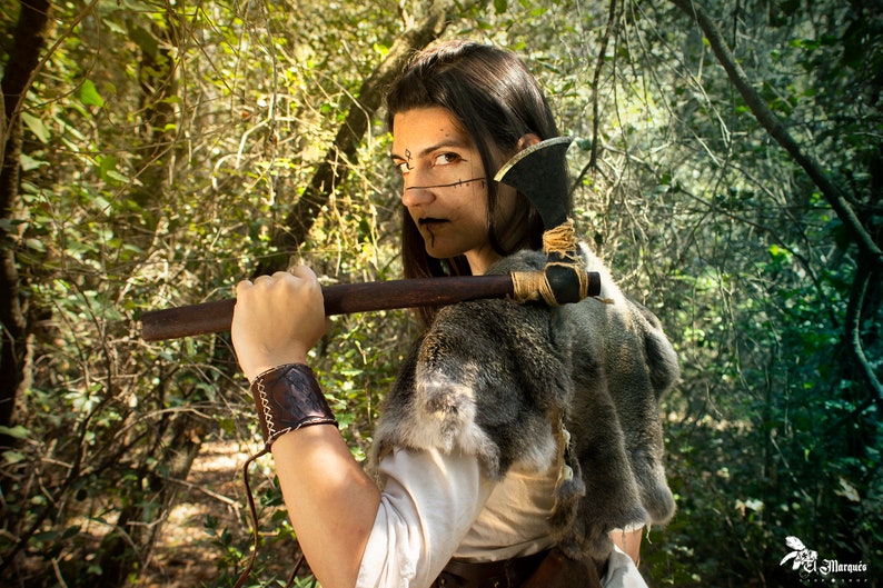 May include: A woman with dark hair and dark makeup is holding a small axe over her shoulder. She is wearing a fur-trimmed garment and a leather wristband. The background is a forest.