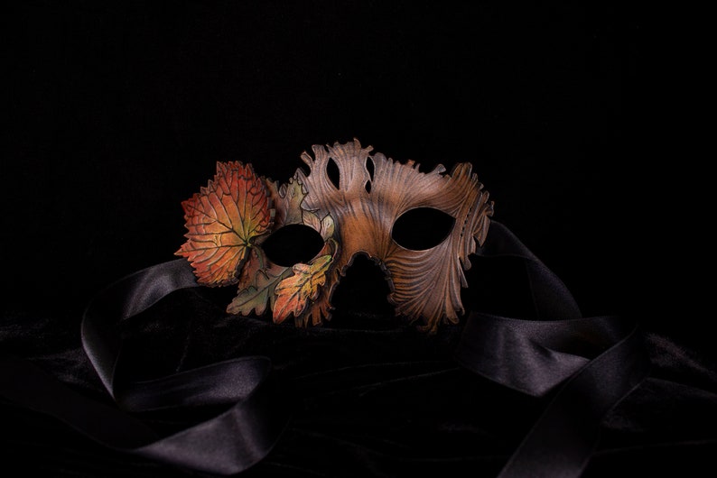 May include: A brown leather masquerade mask with a design of autumn leaves. The mask has a black ribbon attached to it.