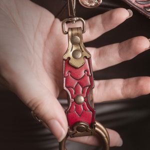 K&ouml;nnte beinhalten: Ein roter Leder-Schl&uuml;sselanh&auml;nger mit goldenen Akzenten und einem Messingring. Der Schl&uuml;sselanh&auml;nger hat ein dekoratives Blumenmuster und ist an einem Metallverschluss befestigt.