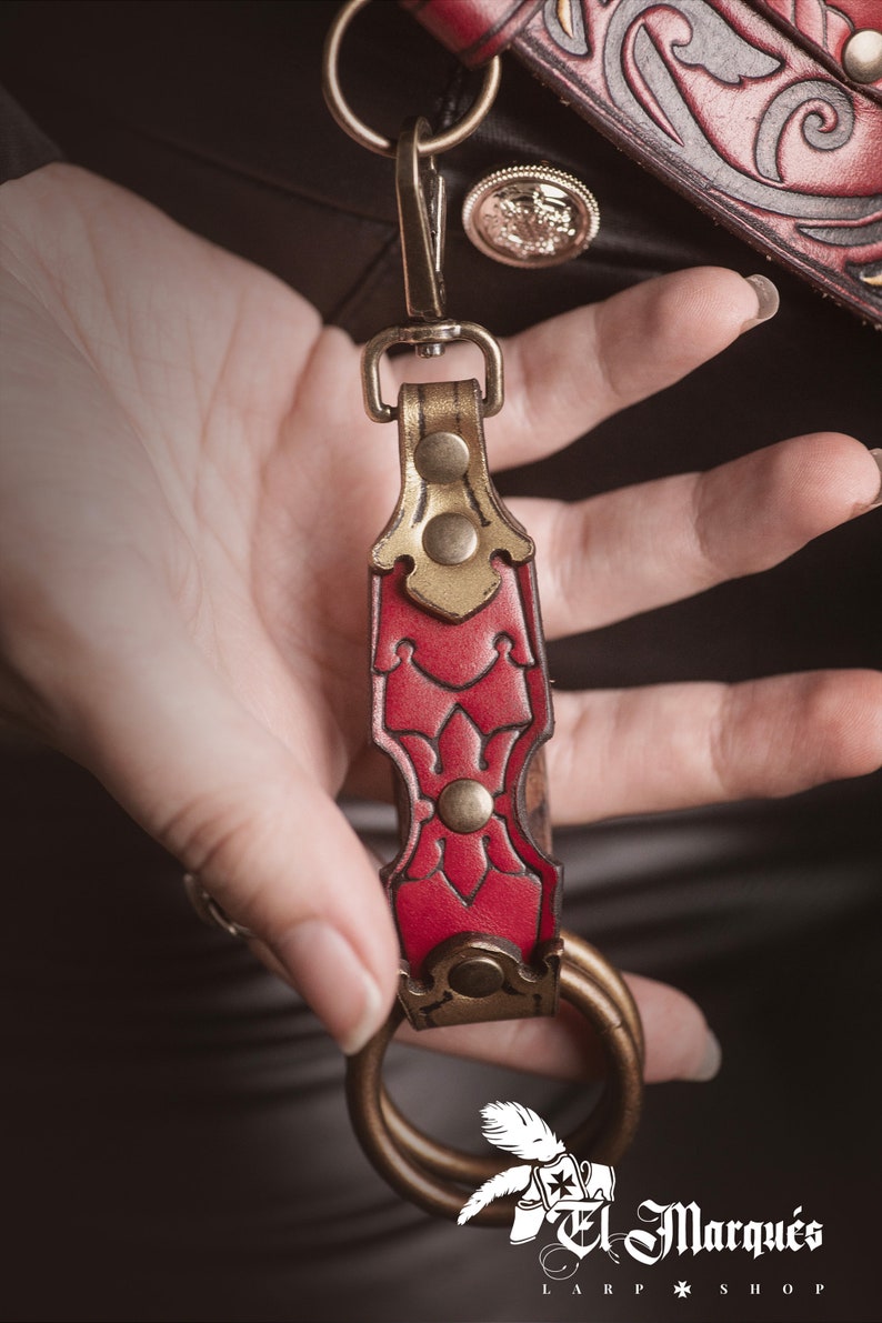 K&ouml;nnte beinhalten: Ein roter Leder-Schl&uuml;sselanh&auml;nger mit goldenen Akzenten und einem Messingring. Der Schl&uuml;sselanh&auml;nger hat ein dekoratives Blumenmuster und ist an einem Metallverschluss befestigt.