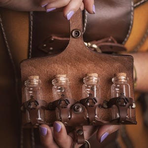 Peut inclure: Une ceinture de potion en cuir marron avec quatre petites fioles en verre, chacune scell&eacute;e avec un bouchon de li&egrave;ge. La ceinture a une boucle pour accrocher et est tenue par une personne avec du vernis &agrave; ongles violet.