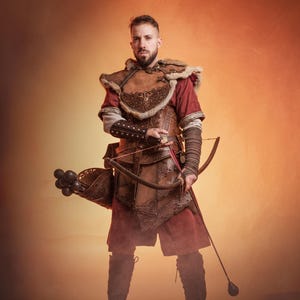 May include: A man wearing a brown leather and fur costume with a bow and arrow. He is standing in front of a warm orange background.