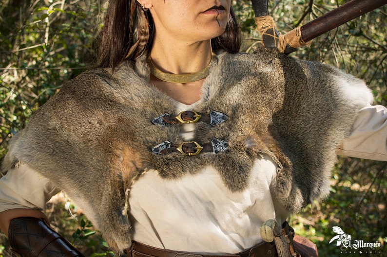May include: A brown fur cape with two brass buckles. The cape is worn over a white shirt and brown leather belt.