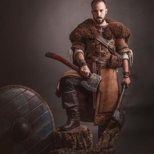 Peut inclure: Une personne en costume de style viking, tenant deux haches. Le costume comprend une tunique en cuir marron, une pi&egrave;ce d'&eacute;paule en fourrure et des bottes noires. Un bouclier en bois est &agrave; gauche. L'image a une esth&eacute;tique sombre et dramatique.