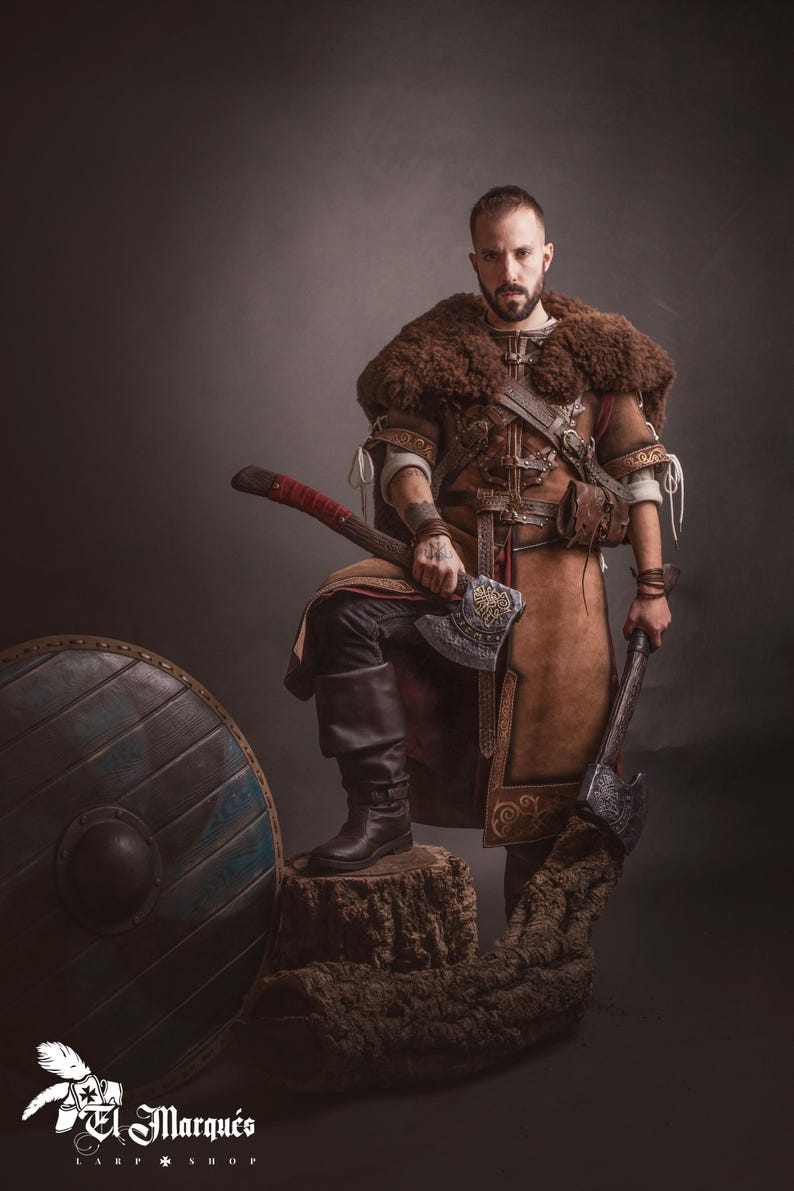 Peut inclure: Une personne en costume de style viking, tenant deux haches. Le costume comprend une tunique en cuir marron, une pi&egrave;ce d'&eacute;paule en fourrure et des bottes noires. Un bouclier en bois est &agrave; gauche. L'image a une esth&eacute;tique sombre et dramatique.