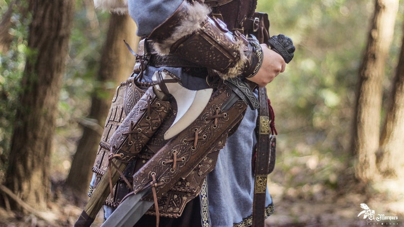 May include: A leather belt with intricate carvings and straps, holding a sword and an axe. The belt is worn over a blue tunic and brown leather armor.