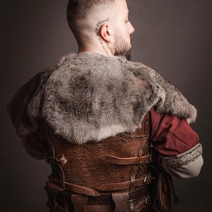 May include: A man wearing a brown leather vest with straps and buckles, a fur-lined cloak, and a red tunic. He is wearing a leather belt with a pouch attached. The vest has intricate designs carved into the leather.