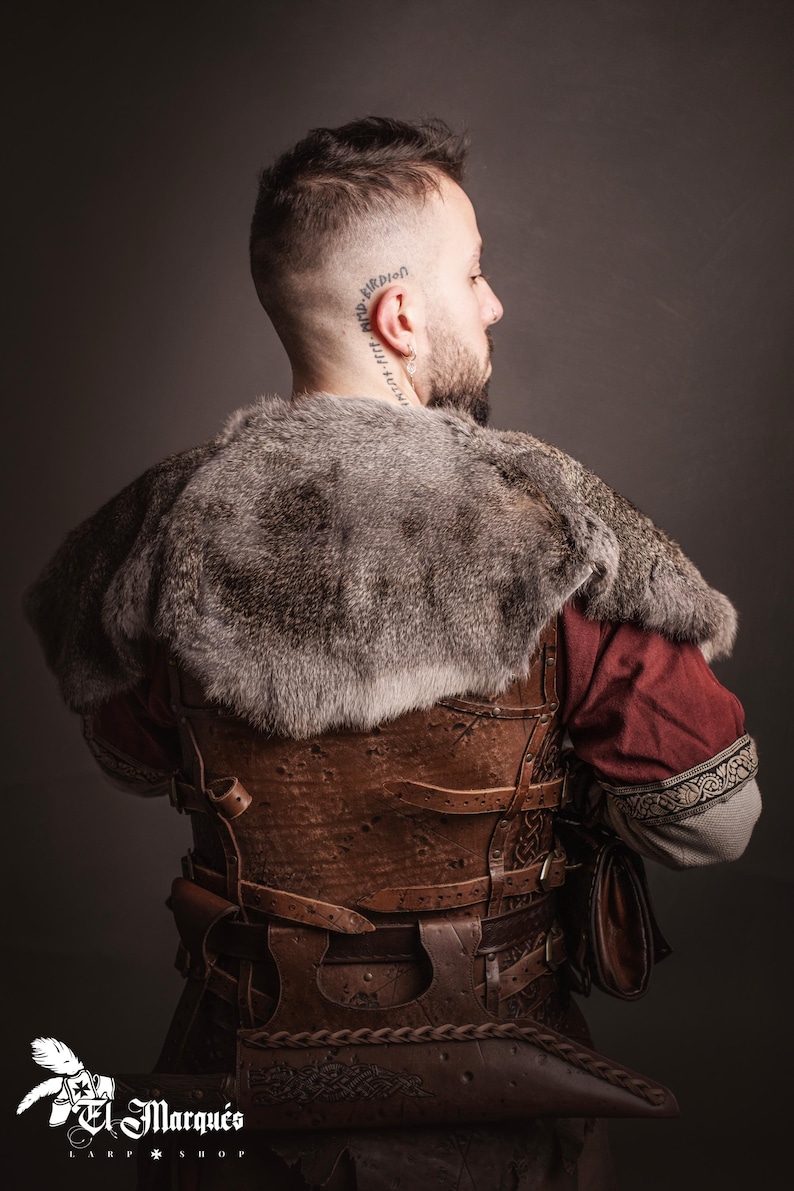 May include: A man wearing a brown leather vest with straps and buckles, a fur-lined cloak, and a red tunic. He is wearing a leather belt with a pouch attached. The vest has intricate designs carved into the leather.