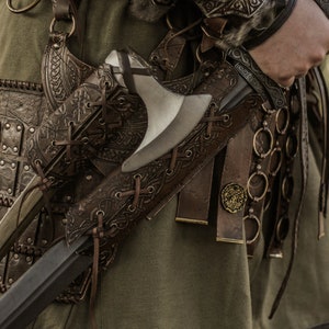 May include: A close-up of a leather scabbard with a metal axe attached. The scabbard is decorated with intricate carvings and has a strap that goes around the waist. The axe has a wooden handle and a metal blade.