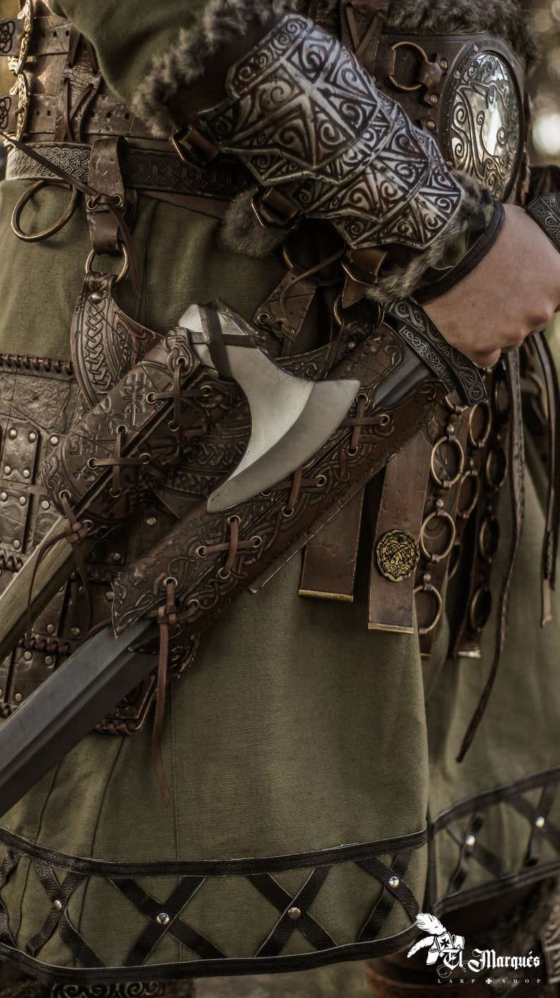 May include: A close-up of a leather scabbard with a metal axe attached. The scabbard is decorated with intricate carvings and has a strap that goes around the waist. The axe has a wooden handle and a metal blade.
