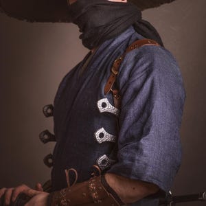 May include: A person wearing a blue kimono with a brown leather strap and silver buckles. They are wearing a black face mask and a wide-brimmed straw hat. The person is holding a sword in their right hand.