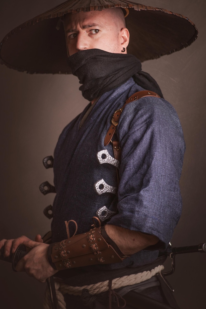 May include: A person wearing a blue kimono with a brown leather strap and silver buckles. They are wearing a black face mask and a wide-brimmed straw hat. The person is holding a sword in their right hand.