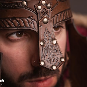 K&ouml;nnte beinhalten: Nahaufnahme eines braunen Lederhelms mit gepr&auml;gten Mustern und Metallnieten. Der Helm hat einen dekorativen Nasenschutz und ist f&uuml;r mittelalterliche oder Fantasy-Themen geeignet. Das Logo "El Marqu&eacute;s" ist sichtbar.