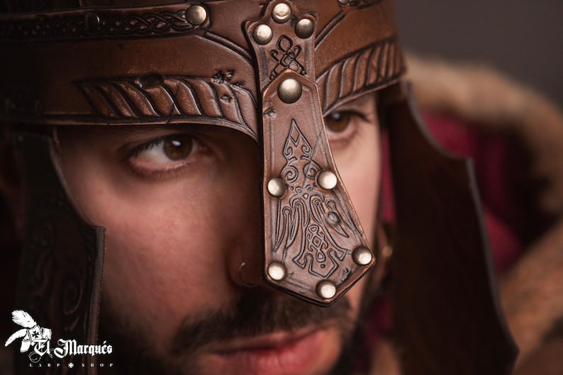 K&ouml;nnte beinhalten: Nahaufnahme eines braunen Lederhelms mit gepr&auml;gten Mustern und Metallnieten. Der Helm hat einen dekorativen Nasenschutz und ist f&uuml;r mittelalterliche oder Fantasy-Themen geeignet. Das Logo "El Marqu&eacute;s" ist sichtbar.