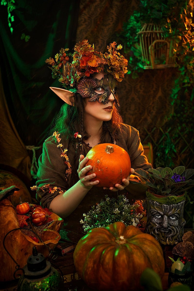 May include: A woman wearing a leafy mask and a crown of autumn leaves holds a small orange pumpkin. She is dressed in a brown tunic with a leafy garland around her neck. The background is a dark green and brown with a birdcage and other fall decorations.