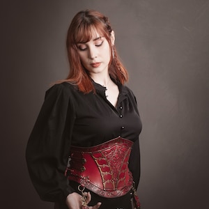 K&ouml;nnte beinhalten: Ein rotes Lederkorsett mit aufwendigen goldenen Details und einem schwarzen Oberteil. Das Korsett hat einen Schn&uuml;rr&uuml;cken und Metallverzierungen. Der Text "El Marqu&eacute;s LARP SHOP" ist in der rechten unteren Ecke sichtbar.