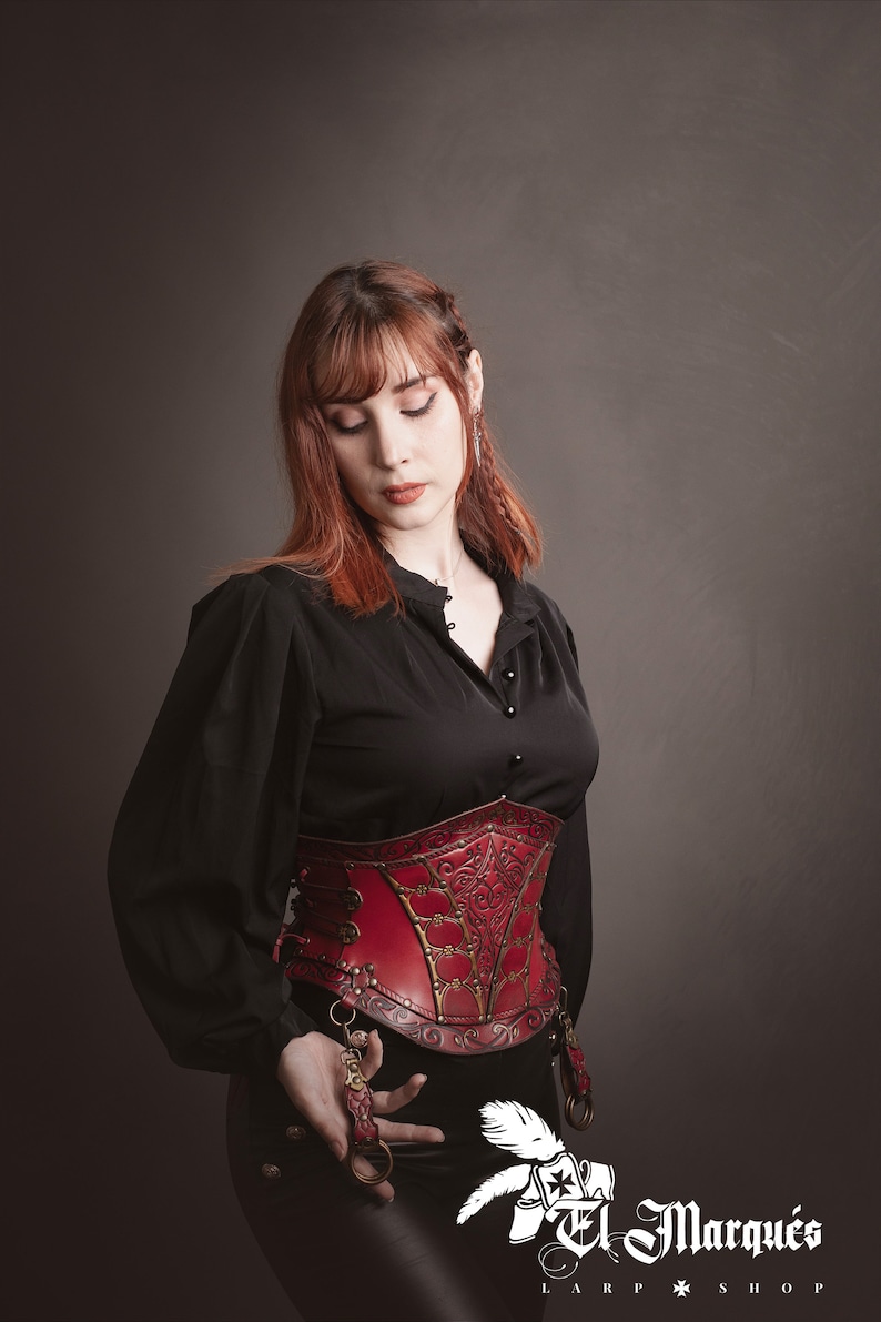 K&ouml;nnte beinhalten: Ein rotes Lederkorsett mit aufwendigen goldenen Details und einem schwarzen Oberteil. Das Korsett hat einen Schn&uuml;rr&uuml;cken und Metallverzierungen. Der Text "El Marqu&eacute;s LARP SHOP" ist in der rechten unteren Ecke sichtbar.