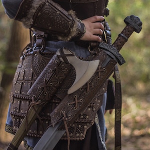 May include: A leather-bound axe and sword sheath with intricate carvings and metal accents. The sheath is worn on a belt with a brown leather strap and metal buckles. The sheath is attached to a brown leather belt with metal buckles.