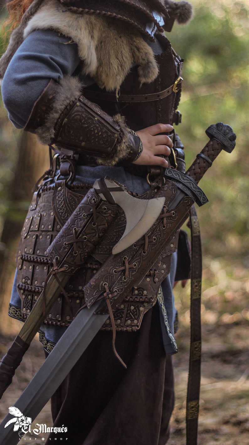 May include: A leather-bound axe and sword sheath with intricate carvings and metal accents. The sheath is worn on a belt with a brown leather strap and metal buckles. The sheath is attached to a brown leather belt with metal buckles.