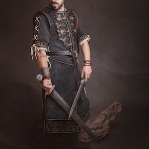 Peut inclure: Un costume m&eacute;di&eacute;val vert fonc&eacute; et noir avec des d&eacute;tails en cuir et des accents dor&eacute;s. La tunique est port&eacute;e avec une ceinture et des bottes assorties. La personne tient deux &eacute;p&eacute;es. L'image comprend le logo El Marqu&eacute;s Larp Shop.