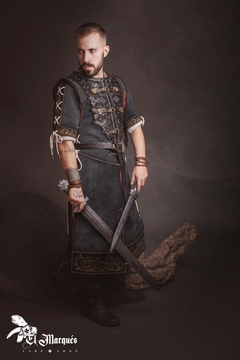 Peut inclure: Un costume m&eacute;di&eacute;val vert fonc&eacute; et noir avec des d&eacute;tails en cuir et des accents dor&eacute;s. La tunique est port&eacute;e avec une ceinture et des bottes assorties. La personne tient deux &eacute;p&eacute;es. L'image comprend le logo El Marqu&eacute;s Larp Shop.