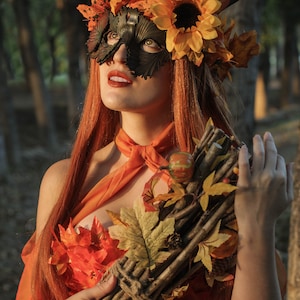 May include: A woman wearing a leafy, autumn-themed mask and crown with a sunflower. She is holding a bundle of sticks and leaves tied together with twine. The woman is wearing a dress made of orange and red leaves.