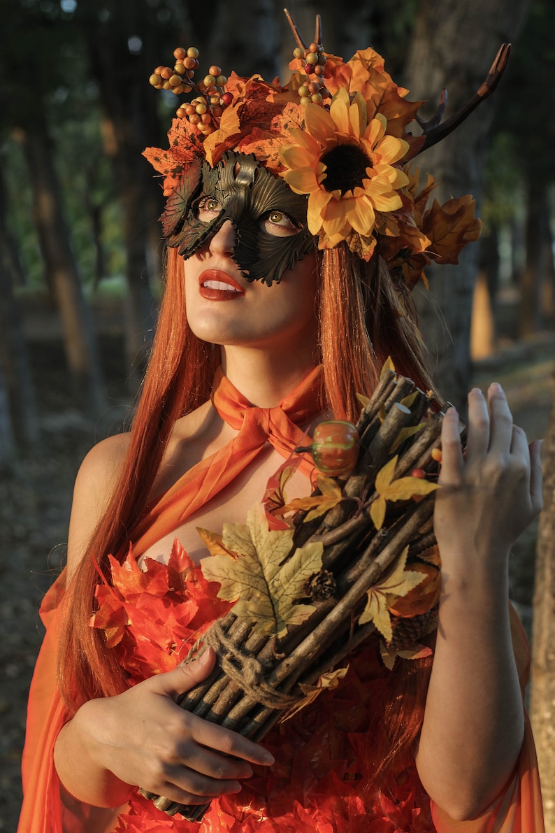 May include: A woman wearing a leafy, autumn-themed mask and crown with a sunflower. She is holding a bundle of sticks and leaves tied together with twine. The woman is wearing a dress made of orange and red leaves.
